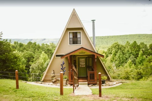 A charming A-frame cabin surrounded by greenery, featuring a wooden porch and stone pathway.