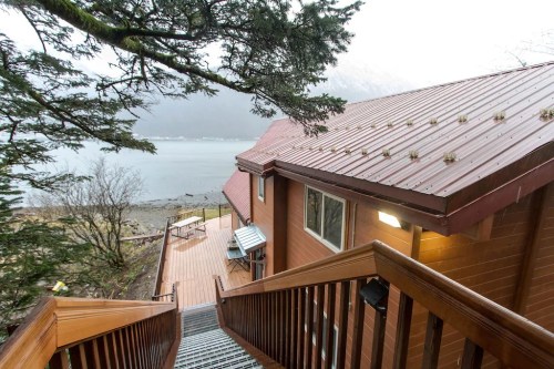 A wooden staircase leads down to a lakeside cabin with a metal roof, surrounded by trees and mountains in the background.