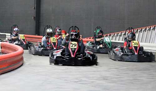 A group of go-karts racing on a track, with drivers wearing helmets and numbered stickers on their karts.