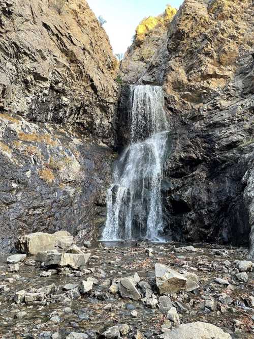 A cascading waterfall flows down rocky cliffs, surrounded by rugged terrain and scattered stones.