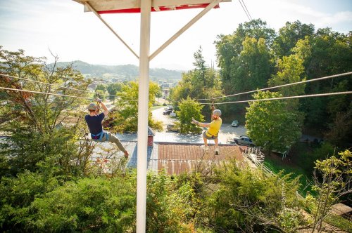 Two people ziplining through a scenic area with trees and buildings below, enjoying a sunny day outdoors.
