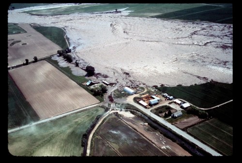Aerial view of a flooded agricultural area, with fields submerged and buildings partially visible amidst the water.