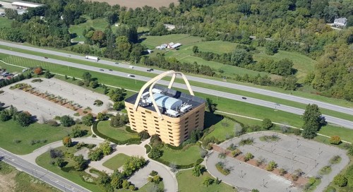 Aerial view of a unique building with large arches, surrounded by greenery and a parking lot near a highway.