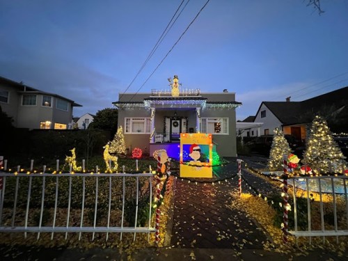 A festive house decorated with colorful Christmas lights, trees, and a large Santa display in the front yard.
