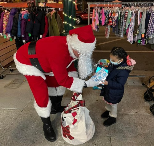 Santa Claus hands a gift to a young girl wearing a mask, surrounded by colorful clothing displays.