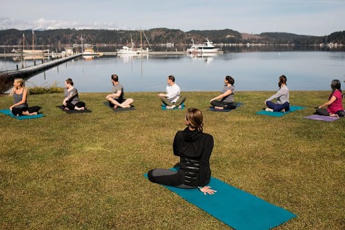 A group of people practicing yoga on mats by a calm lake, with boats and mountains in the background.