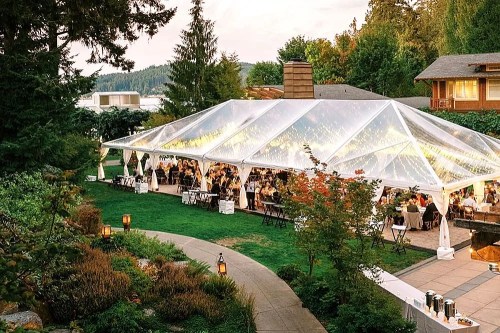 A large outdoor tent filled with guests dining, surrounded by greenery and trees, with soft lighting in the evening.