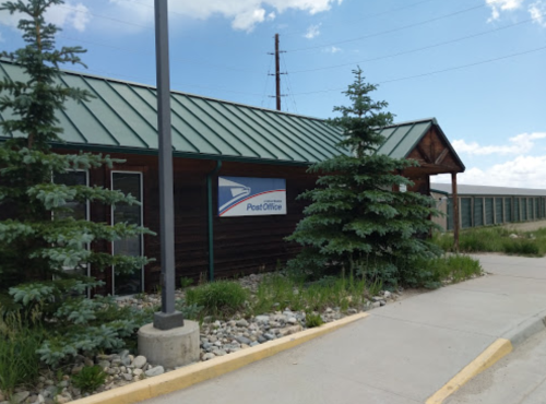 A small post office building with a green metal roof, surrounded by trees and a clear blue sky.