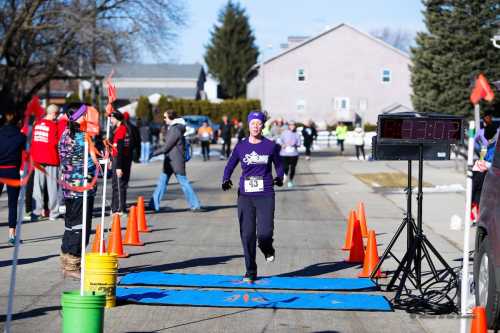 A runner crosses the finish line at a race, with spectators and volunteers cheering in the background.