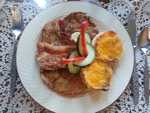 A plate of pancakes with bacon, accompanied by baked egg cups topped with cheese and garnished with cucumber and bell pepper.