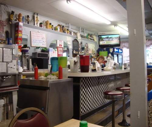 A diner interior featuring a counter with colorful drink cups, condiments, and a staff member preparing food.