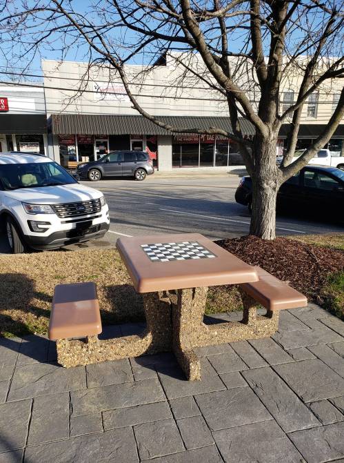 A picnic table with a checkerboard top sits on a paved area near parked cars and a tree.