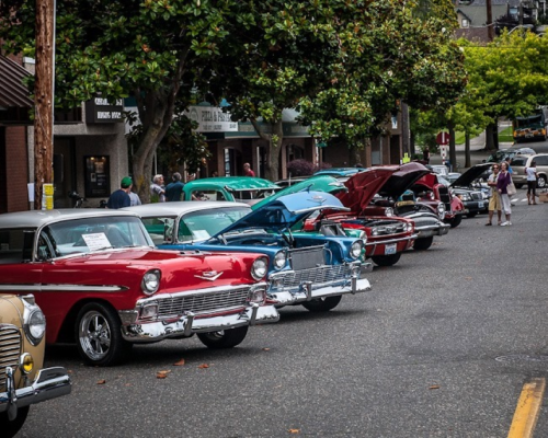 A row of classic cars in various colors parked along a street, with people admiring them and trees in the background.