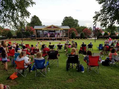 A crowd sits in folding chairs, enjoying a live performance on an outdoor stage surrounded by trees and greenery.