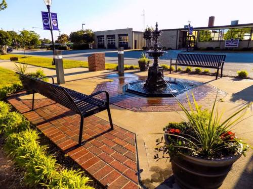 A decorative fountain surrounded by benches and planters in a landscaped area, with a street and buildings in the background.