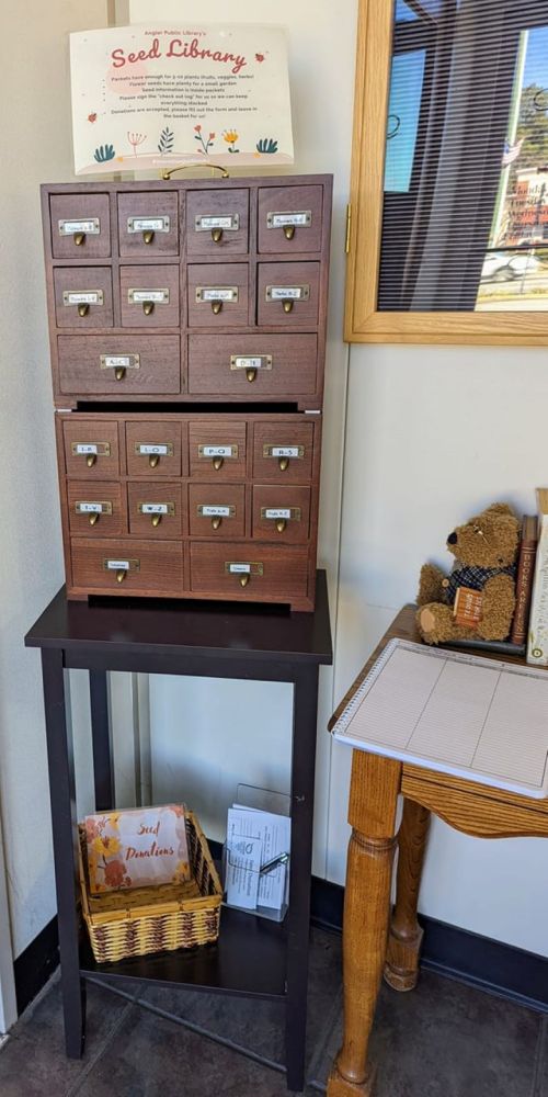 A wooden seed library cabinet with labeled drawers, next to a table with a basket and a stuffed bear.