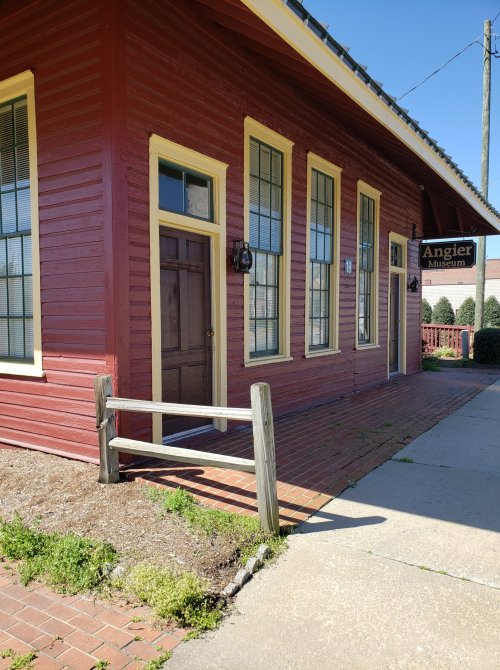 A red building with yellow trim, featuring large windows and a sign for the Angier Museum, surrounded by greenery.