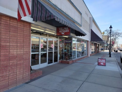 Exterior of Village Antiques shop with a striped awning, American flag, and sidewalk sign in a shopping area.