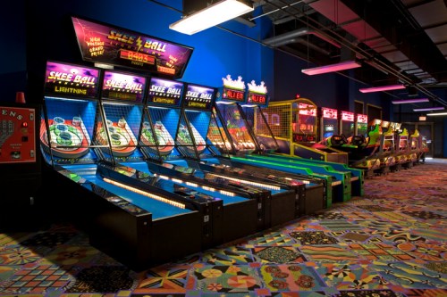 A vibrant arcade featuring multiple skee-ball machines and colorful flooring, illuminated by bright lights.
