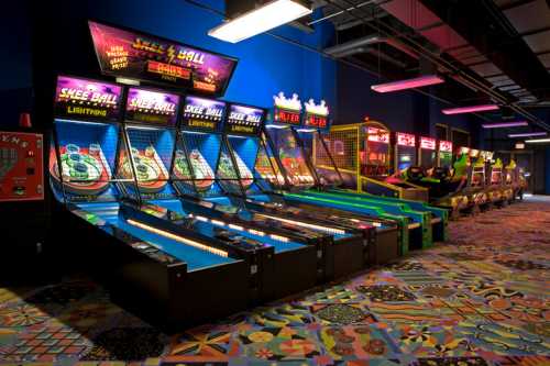 A vibrant arcade featuring multiple skee-ball machines and colorful flooring, illuminated by bright lights.