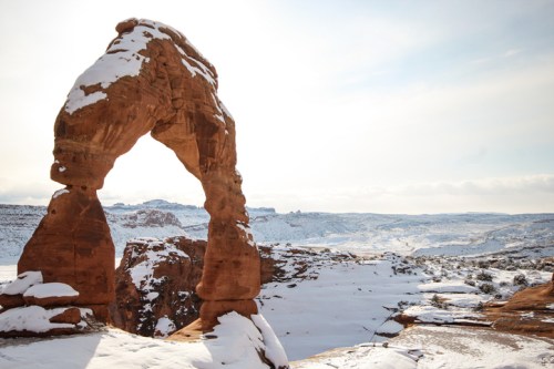 A snow-covered rock arch stands against a bright sky, surrounded by a winter landscape of hills and valleys.