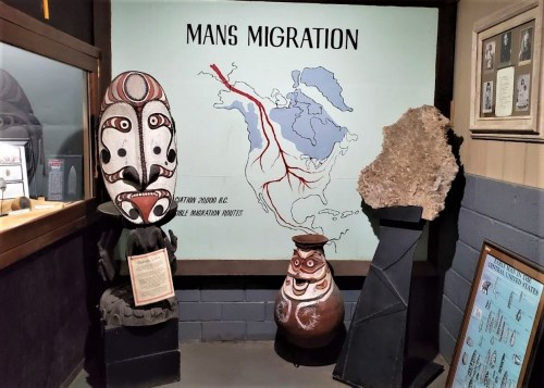 Display featuring artifacts related to human migration, with a map and decorative masks in a museum setting.