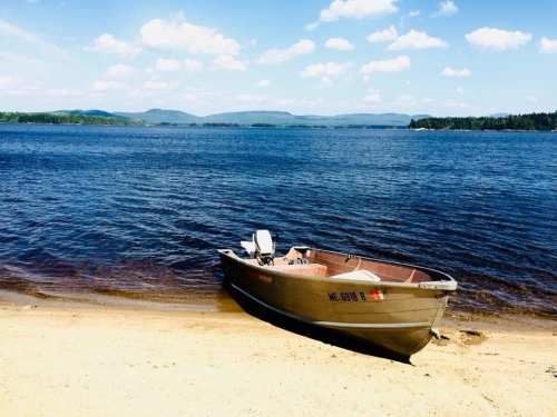 A small boat rests on a sandy shore by a calm lake, with mountains and blue skies in the background.