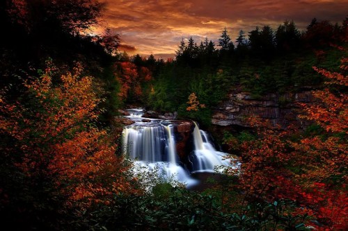 A serene waterfall surrounded by vibrant autumn foliage and a dramatic sunset sky.