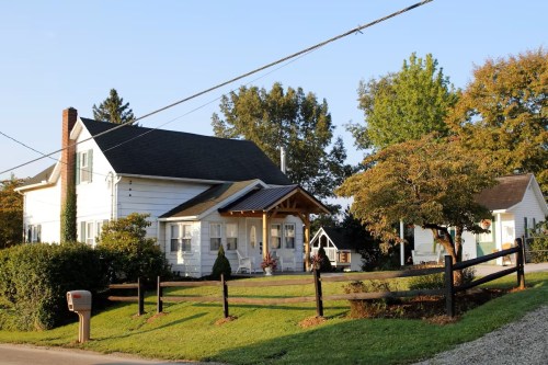 A charming white house with a porch, surrounded by greenery and trees, on a sunny day. A mailbox is visible in the foreground.