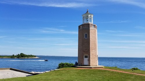 A tall lighthouse stands on a grassy shore, overlooking a calm blue sea under a clear sky.