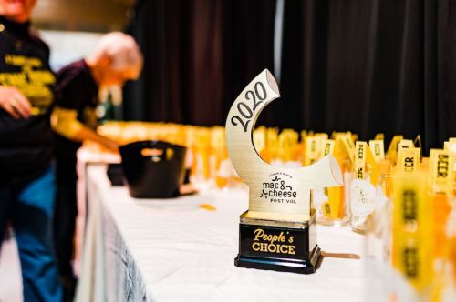 A trophy labeled "2020 People's Choice" stands on a table with mac and cheese festival awards in the background.