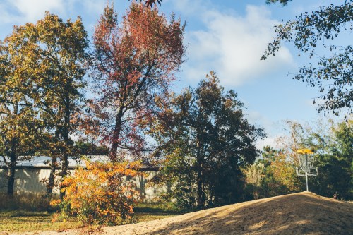 A disc golf basket on a hill surrounded by trees with autumn foliage under a partly cloudy sky.