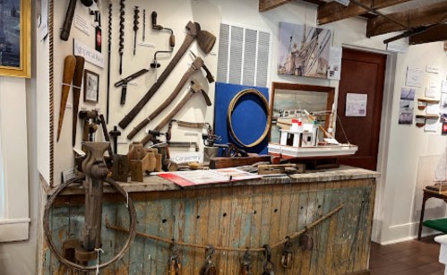 A display of vintage carpentry tools and a model boat on a rustic wooden table in a museum setting.