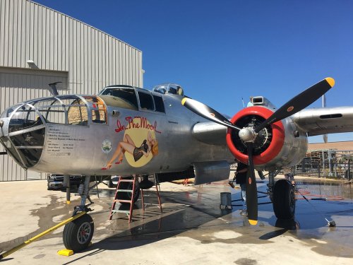 A vintage bomber aircraft with a pin-up girl mural, parked near a hangar, showcasing its shiny metal exterior.