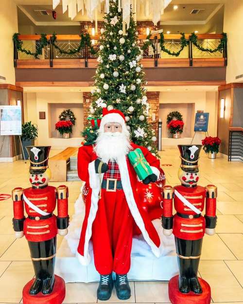 A festive scene featuring a Santa figure with gifts, flanked by two toy soldiers, in front of a decorated Christmas tree.
