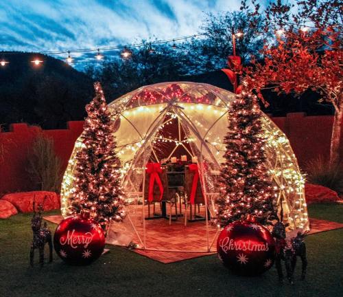 A festive outdoor igloo decorated with lights, Christmas trees, and red ornaments, set against a twilight sky.