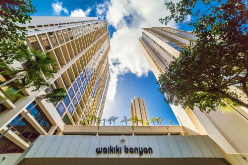 View of the Waikiki Banyan building, showcasing tall towers against a blue sky with scattered clouds.