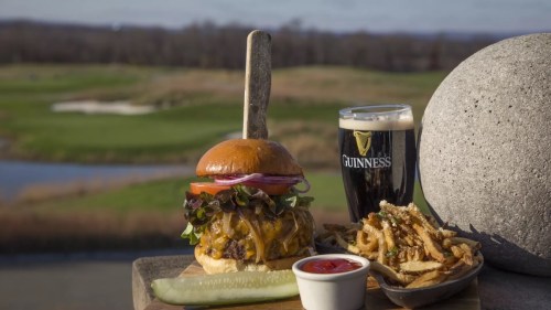 A gourmet cheeseburger with toppings, fries, a pickle, and a pint of Guinness, set against a scenic golf course backdrop.