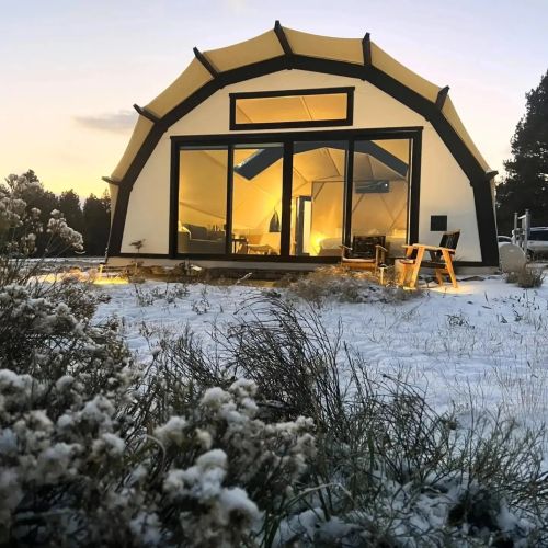 A modern tent-like structure in a snowy landscape, with warm light glowing from inside at sunset.
