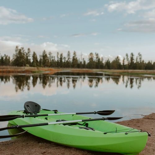 Two green kayaks are parked on a sandy shore beside a calm, reflective lake surrounded by trees.