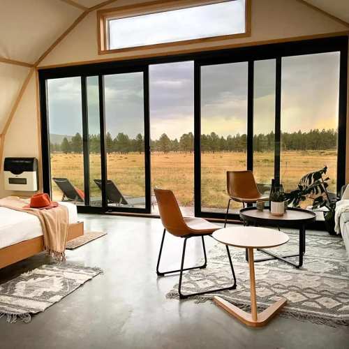 A cozy interior with a bed, chairs, and a table, featuring large windows overlooking a scenic outdoor view.