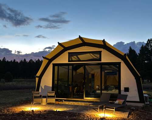A modern tent-like structure with large windows, illuminated at dusk, surrounded by trees and a clear sky.