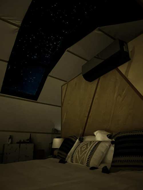 Cozy bedroom with a starry sky visible through a skylight, featuring a wooden wall and decorative pillows.