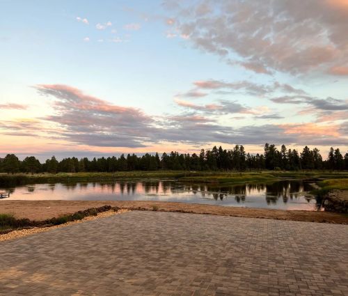 A serene landscape featuring a calm lake, lush greenery, and a colorful sunset sky with scattered clouds.