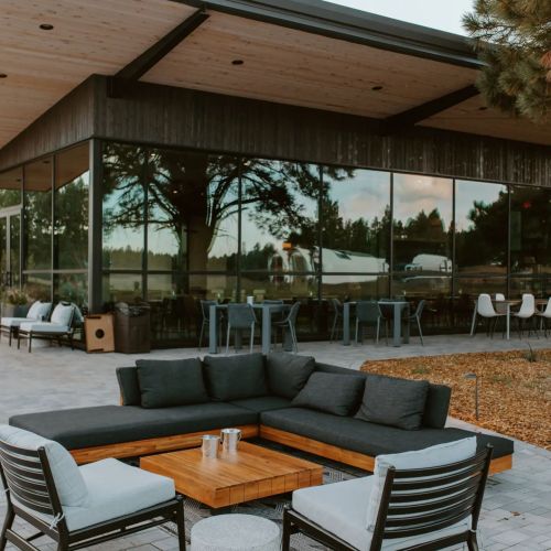 A modern outdoor seating area with a sectional sofa and coffee table, surrounded by large windows and greenery.