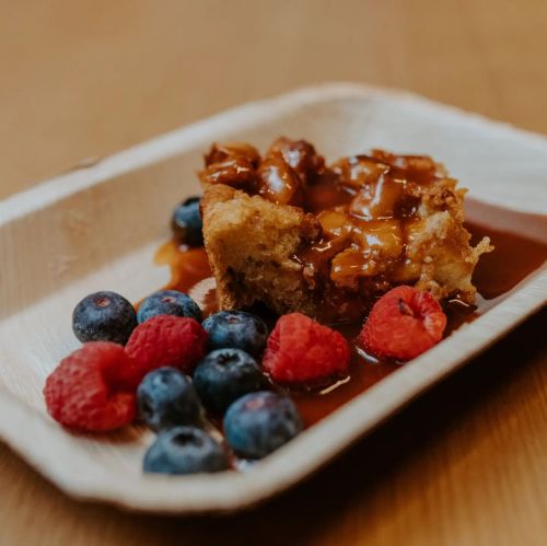 A slice of cake drizzled with caramel sauce, served with fresh blueberries and raspberries on a wooden plate.