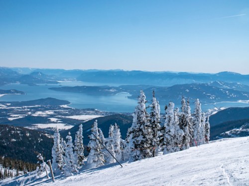 A snowy mountain landscape with frosted trees overlooking a serene lake and distant hills under a clear blue sky.