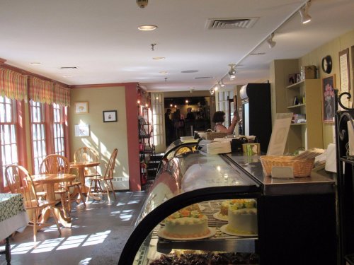 Cozy café interior with tables, a display case of desserts, and sunlight streaming through large windows.