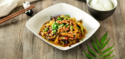A plate of stir-fried noodles with vegetables and meat, served with a bowl of rice and chopsticks on a wooden table.