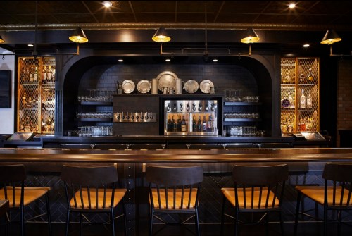 A stylish bar with a dark wood counter, shelves of glassware, and decorative plates, featuring empty chairs in front.